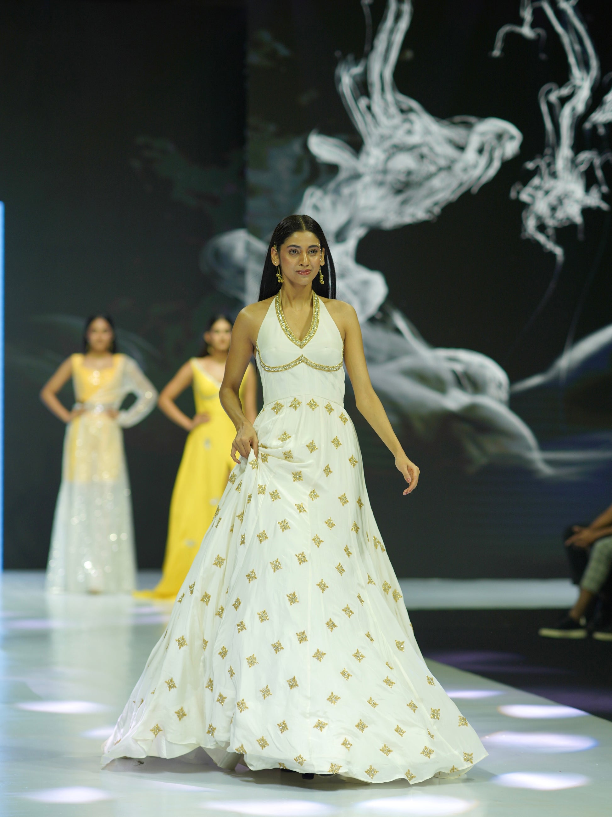 Model walking on a runway wearing a white dress with gold patterns, with other models in the background.
