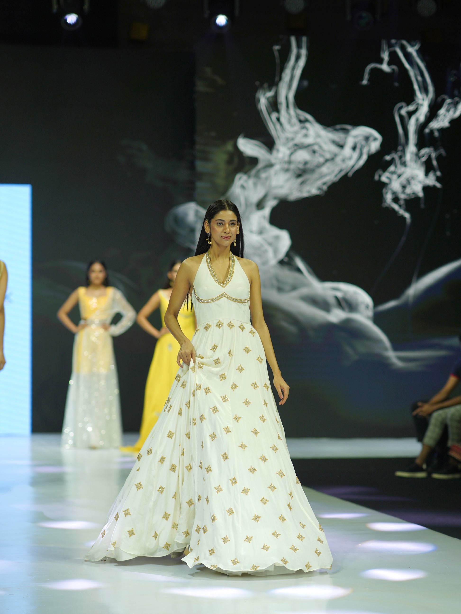 Model walking on a fashion show runway wearing a white dress with gold patterns.