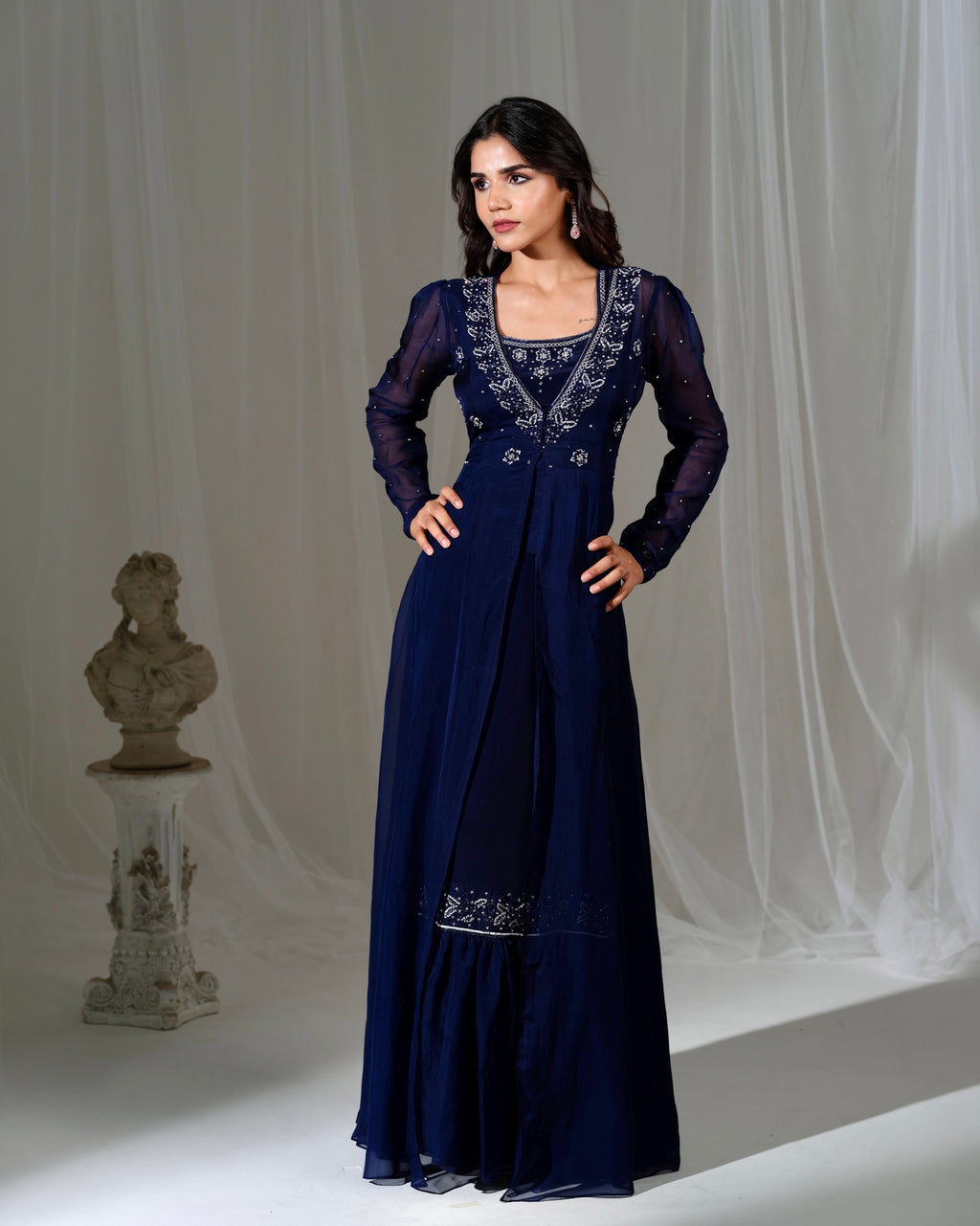 Woman wearing a long navy blue dress with intricate embroidery against a white curtain background