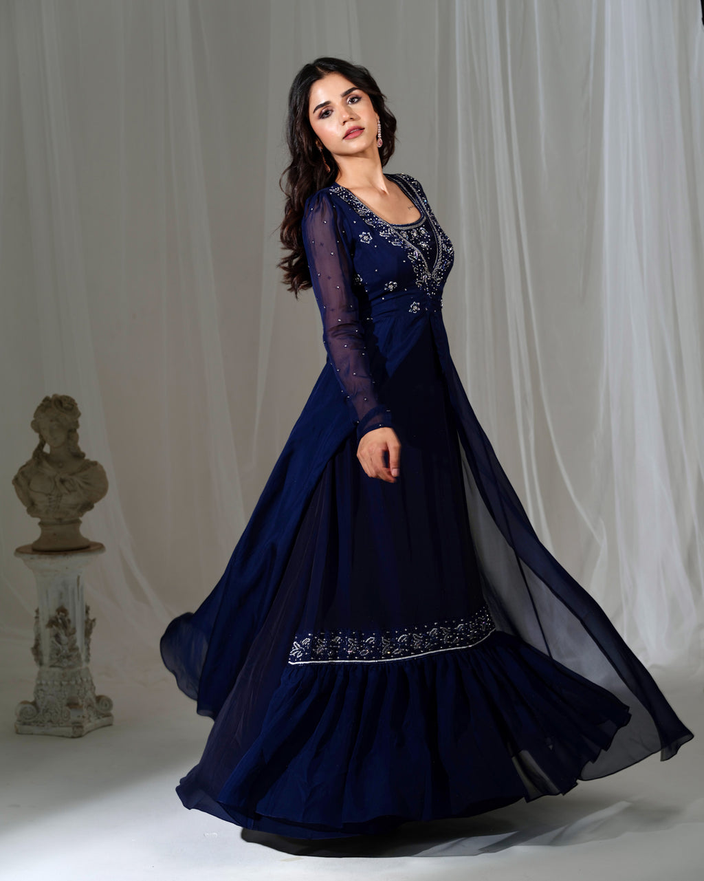 Woman wearing a dark blue embroidered dress against a white curtain background