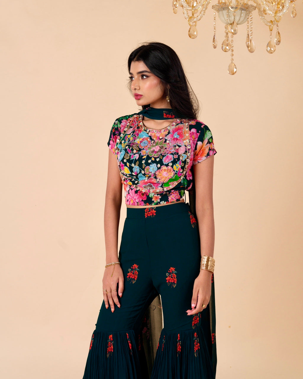 Woman wearing a floral top and dark green pants with embroidery, standing against a beige wall with a chandelier.