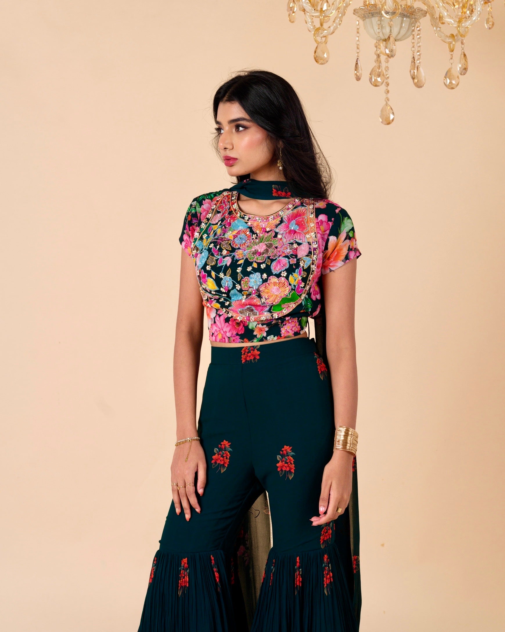 Woman wearing a floral top and dark green pants with embroidery, standing against a beige wall with a chandelier.