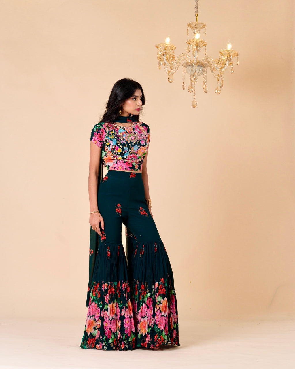 Woman wearing a floral top and dark green pants with a beige background and chandelier.