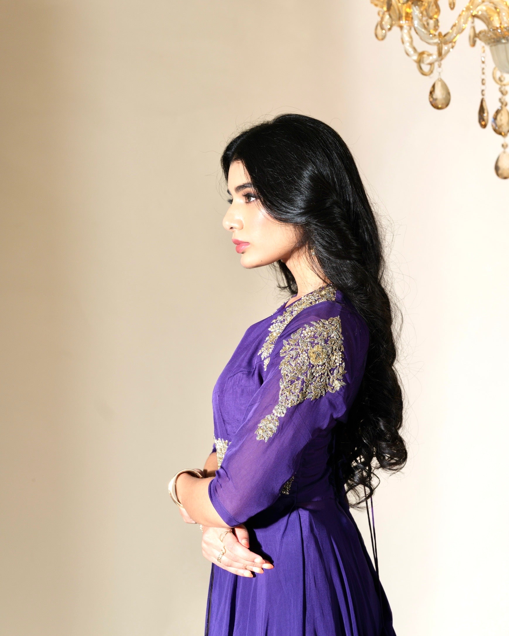 Woman wearing a purple dress with gold embroidery against a beige wall.
