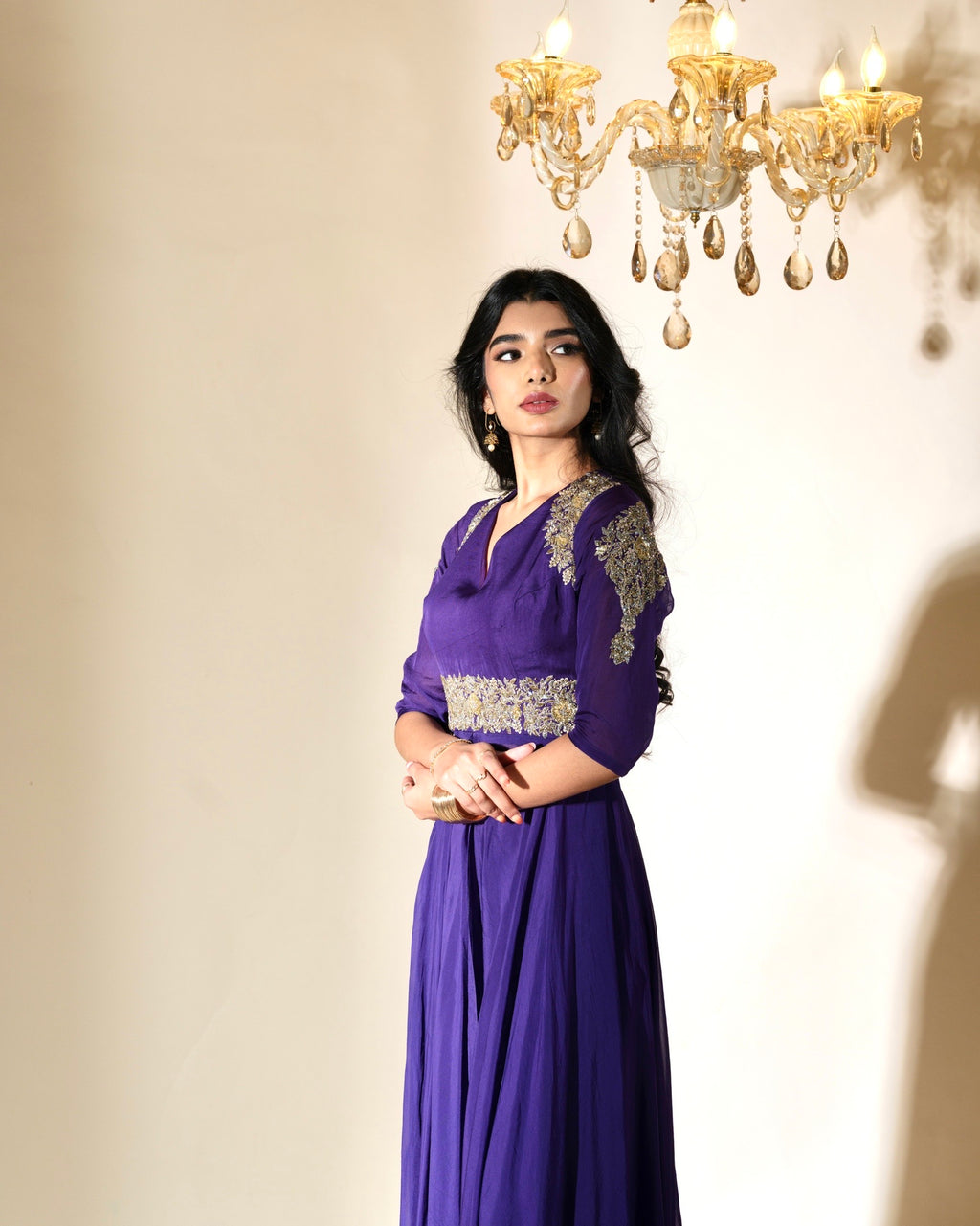 Woman wearing a purple dress with gold embroidery in a softly lit room with a chandelier.