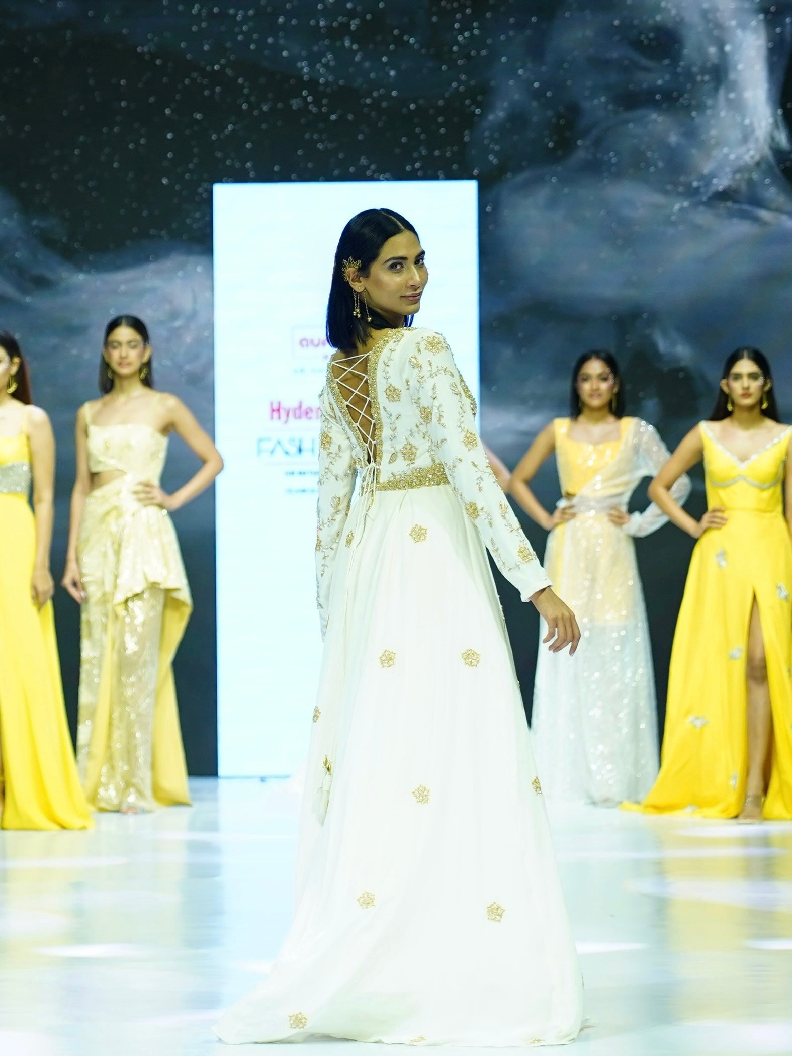 Model on a catwalk wearing a white and gold outfit with other models in yellow outfits in the background.