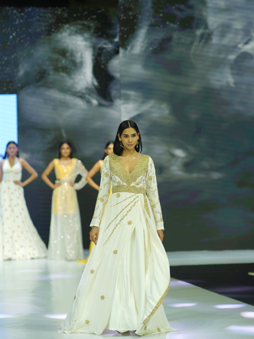 Model wearing a white saree with gold details on a fashion show runway.