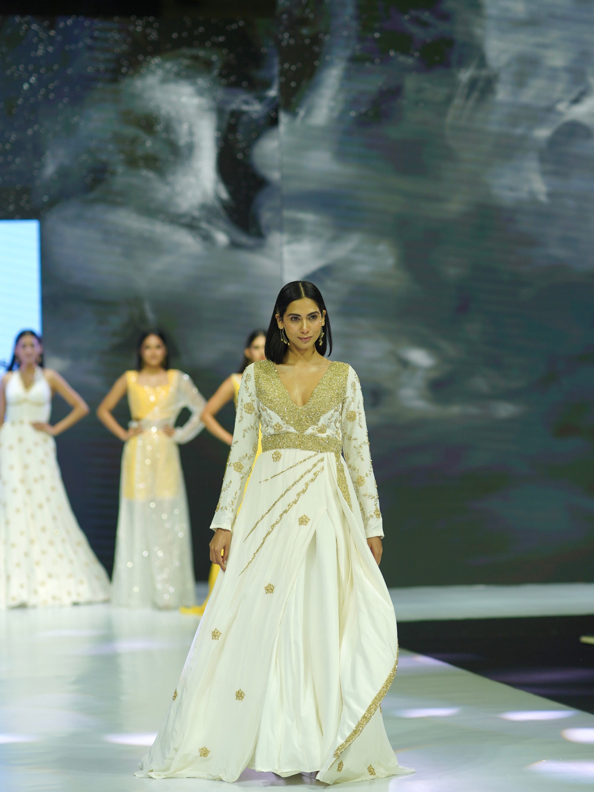 Model wearing a white saree with gold details on a fashion show runway.