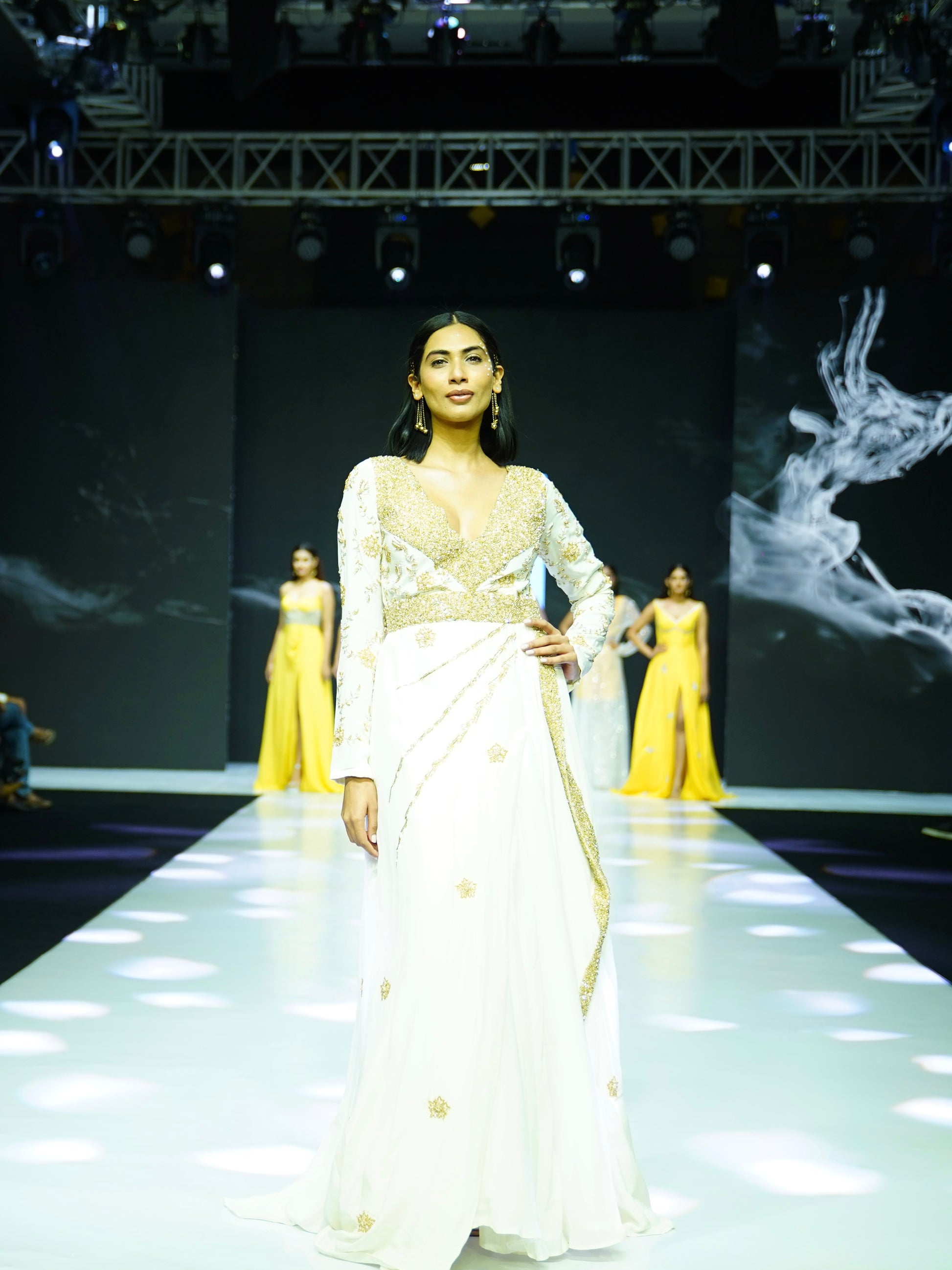 Model walking on a fashion show runway wearing a white and gold dress.