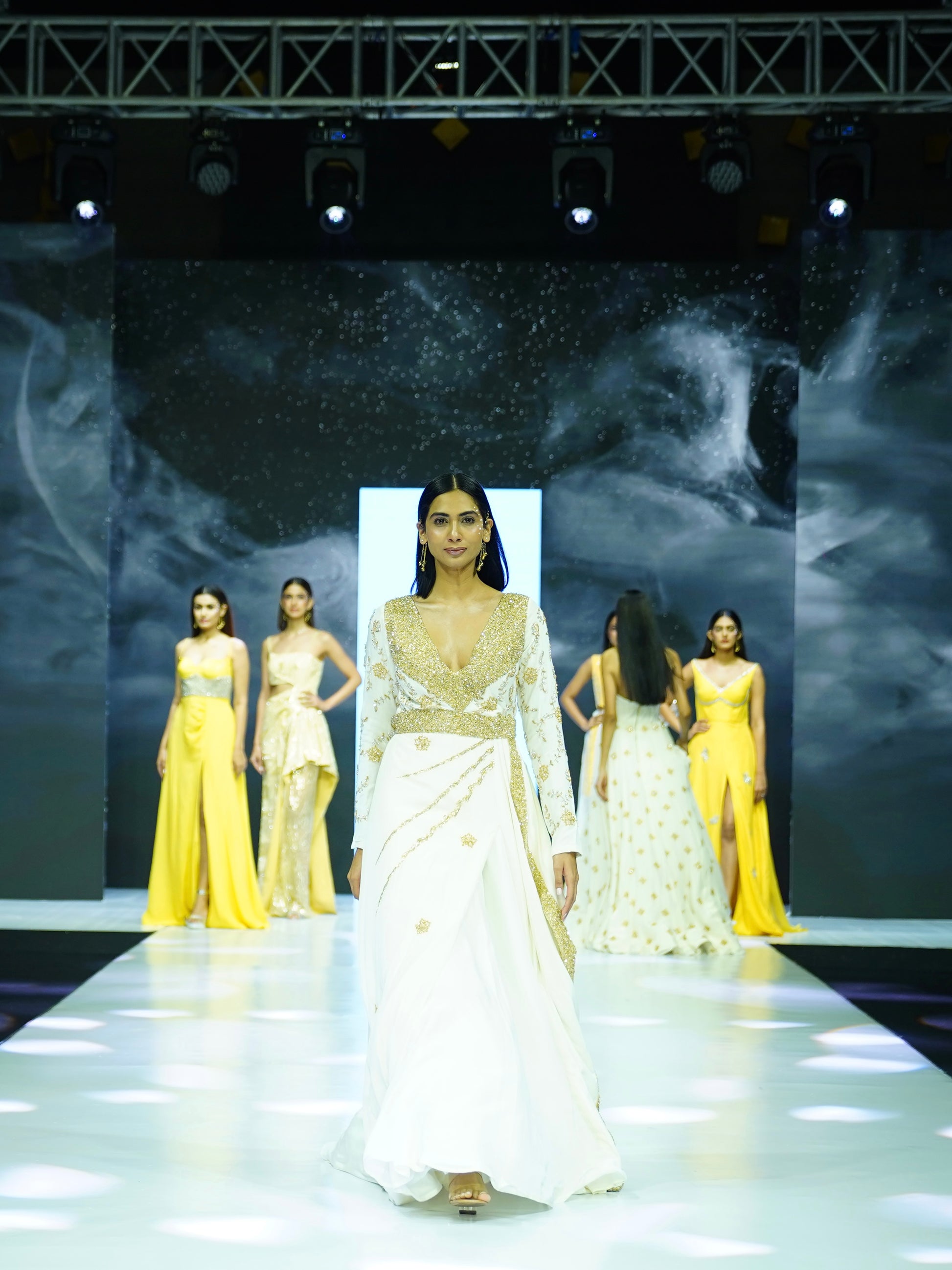 Model walking on a runway wearing a white dress with other models in yellow dresses in the background.