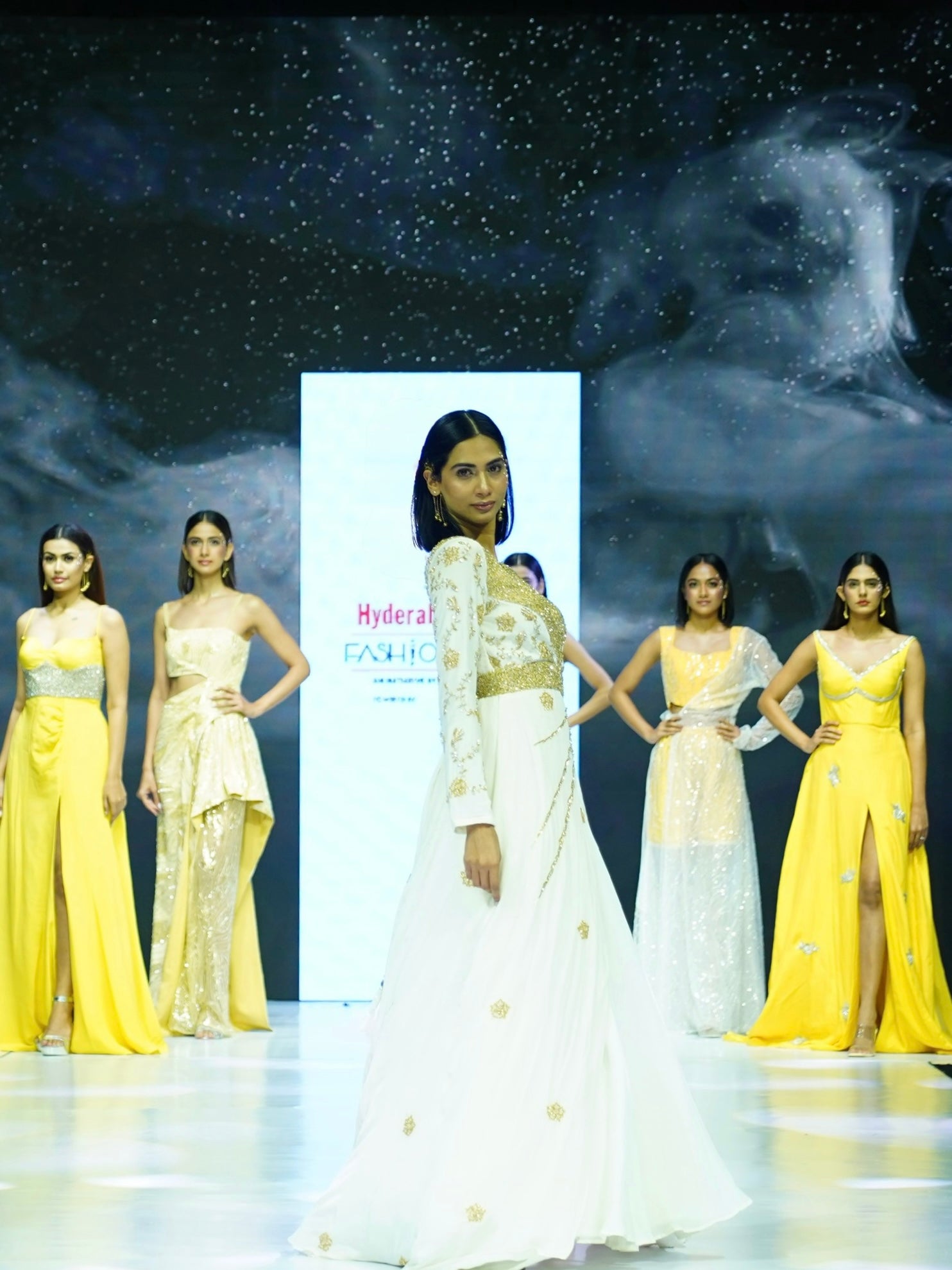 Model in a white and gold dress on a runway with a 'Hyderabad Fashion Week' banner in the background.