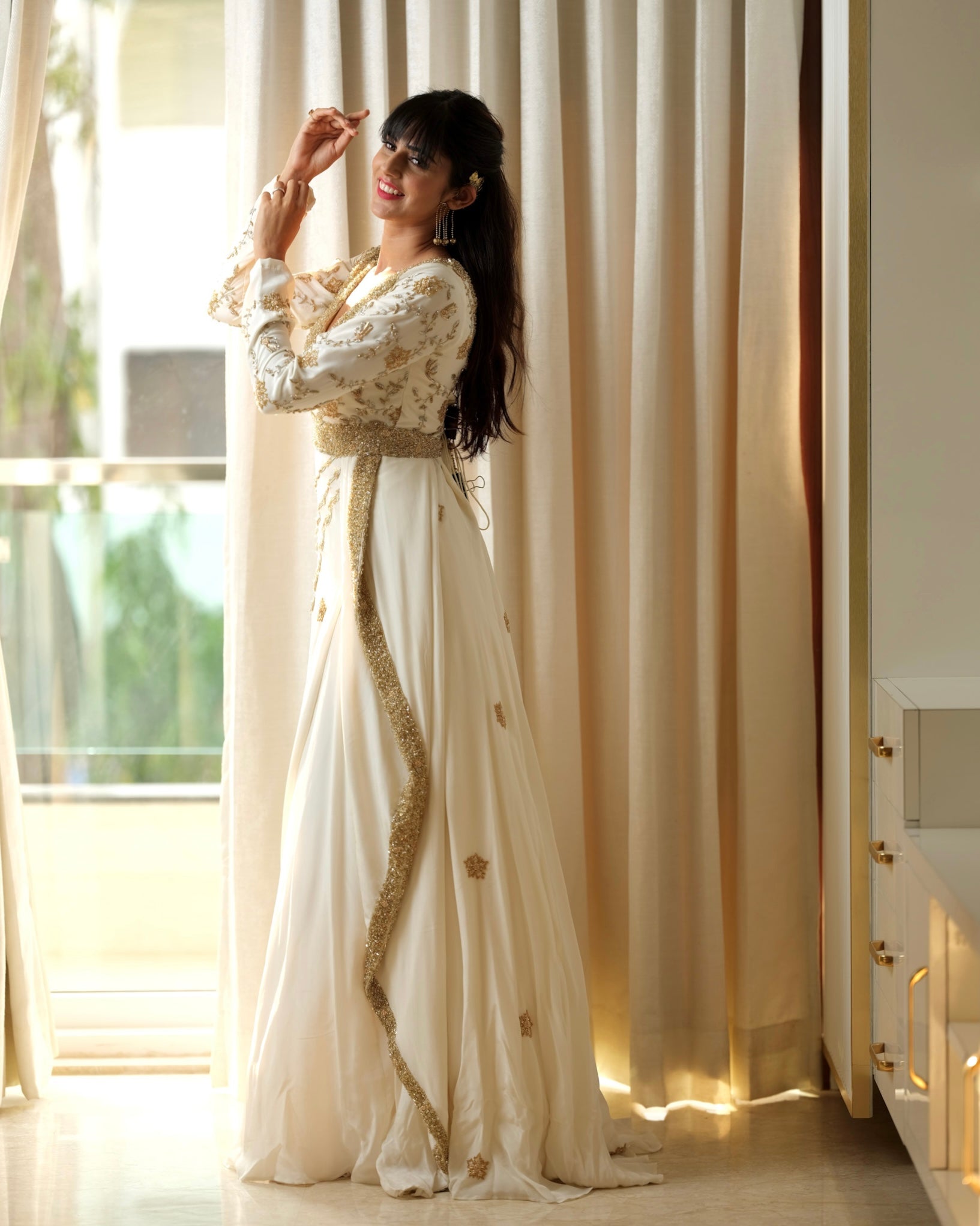 Woman in a white and gold traditional outfit standing in a bright room with large windows.