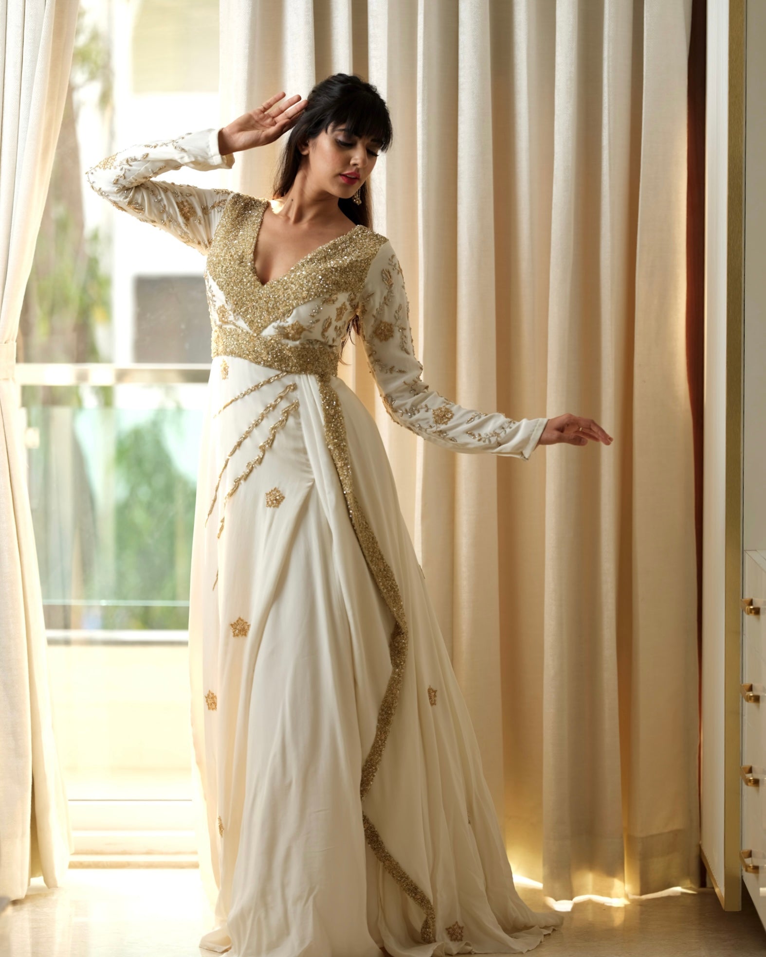 Woman in a white and gold saree standing in a bright room with large windows.