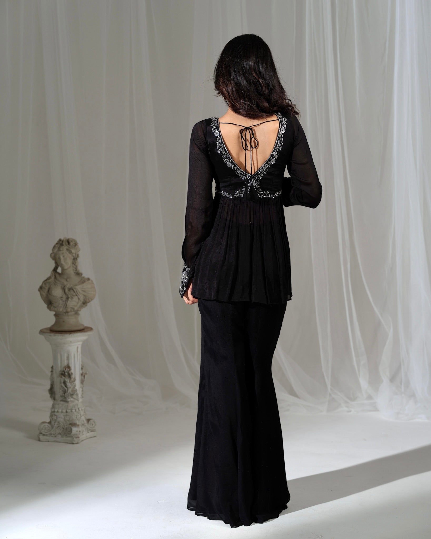 Woman wearing a black evening gown with intricate back design against a white curtain background.